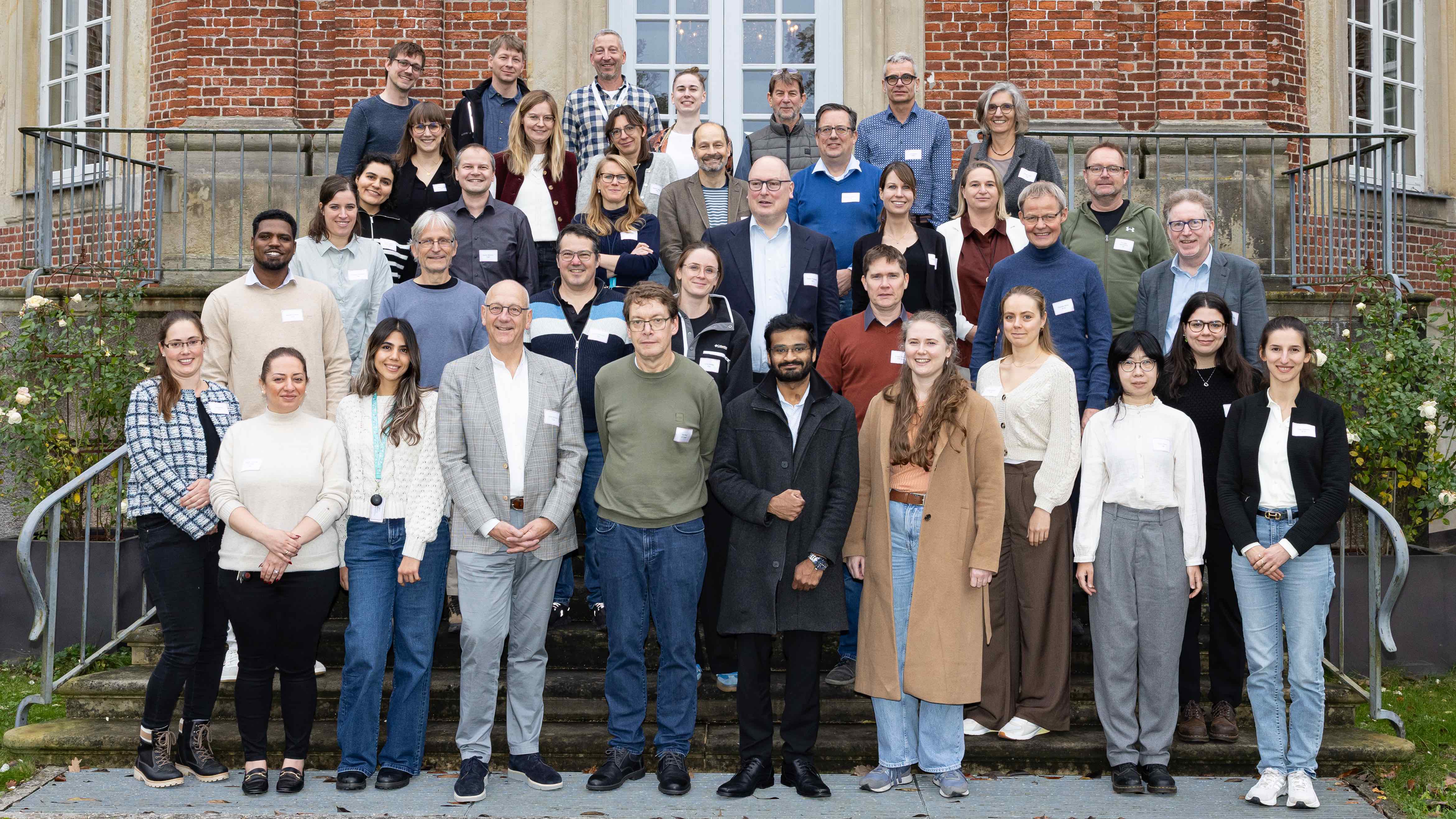 A large group of people are standing on the stone steps in front of a historic red brick building for a group photo.