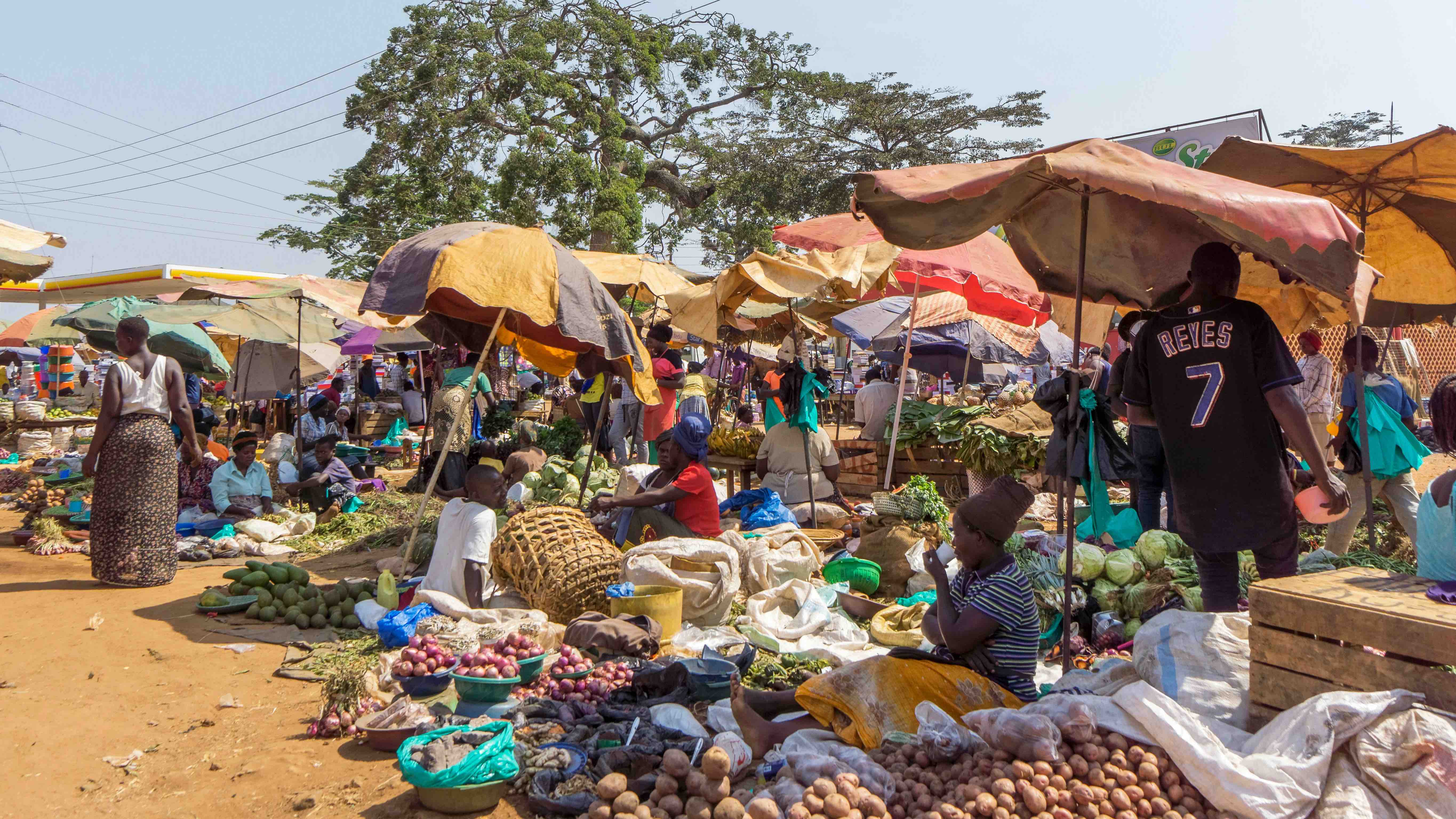 Ein Markt in Afrika unter freiem Himmel. Verkäuferinnen und Verkäufer haben ihre Waren auf dem Boden ausgebreitet. Die Stände sind durch bunte Sonnenschirme vor der Sonne geschützt.